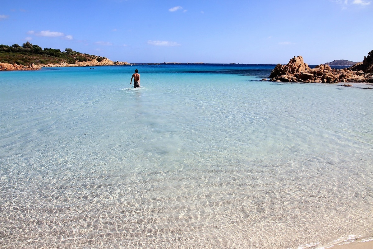 A serene beach scene depicts clear turquoise waters gently lapping against the sandy shoreline. A person wades into the shallow water, with rocky formations in the background and a distant coastline visible under a clear blue sky.