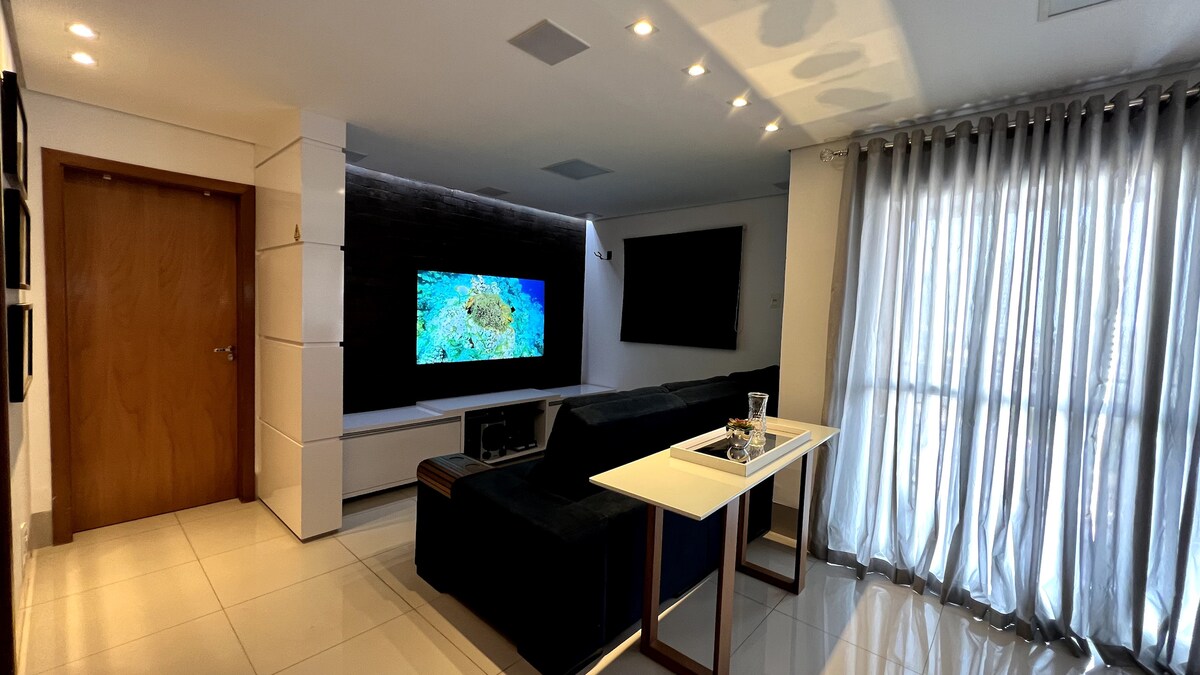 A modern living area is shown, featuring a spacious sofa facing a large wall-mounted TV. Bright lighting enhances the glossy tile floor, while sheer curtains frame a wide window, allowing natural light to filter in. A small side table rests beside the sofa.