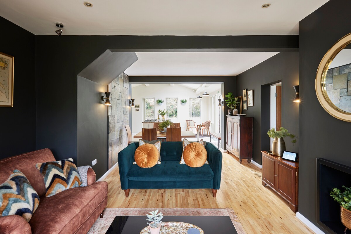 A living area showcases a rich blend of textures, featuring a deep blue sofa with orange accent pillows and a cozy brown couch. Natural light floods in from the adjoining dining space, highlighting the hardwood flooring and stone wall accents.
