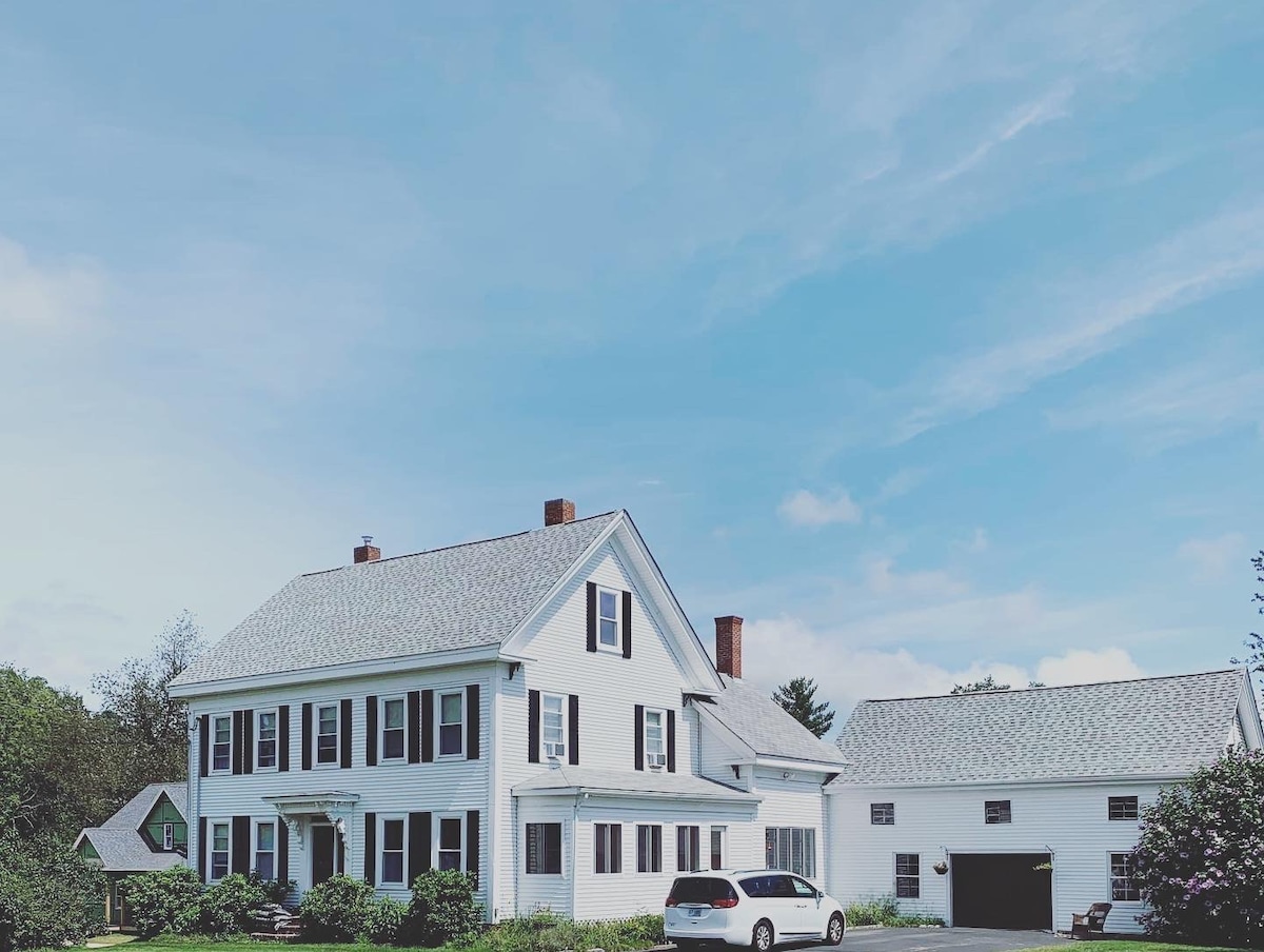 The modern farmhouse is showcased in this image, featuring a classic white exterior with black shutters. A spacious driveway allows for ample parking. The large structure is accompanied by a garage, with lush greenery and a clear blue sky providing a serene backdrop.