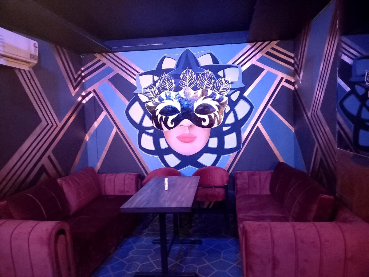 A stylish lounge area is highlighted by plush red seating arranged around a dark wooden table. The walls feature an intricate geometric design in blue and gold tones. Ambient lighting creates a relaxed atmosphere, enhancing the modern decor.