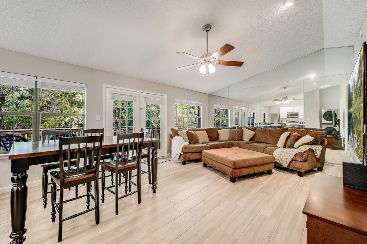 A spacious living area features a large sectional sofa and a dining table with four chairs. Natural light enters through multiple windows, providing views of the wooded surroundings. The open layout leads to the kitchen area, enhancing a sense of connectivity.