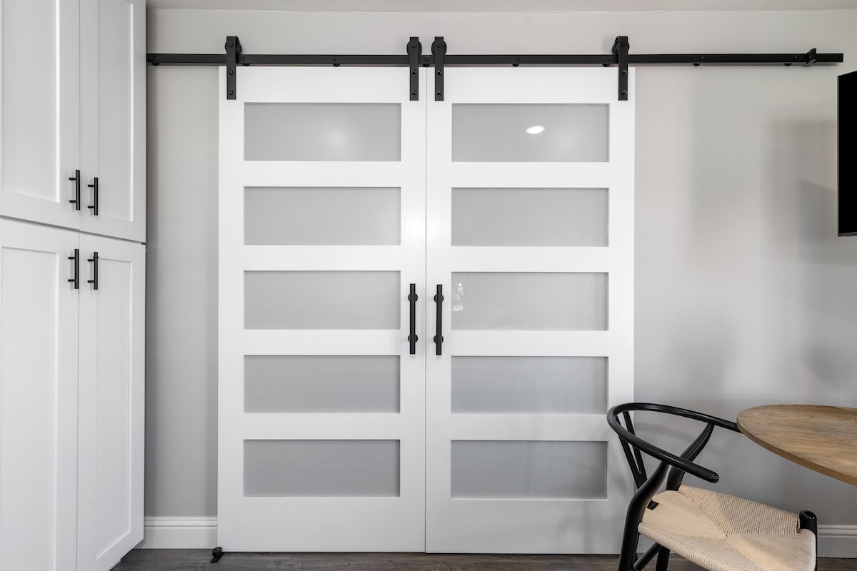 Two modern barn doors, featuring frosted glass panels, provide a stylish separation between the living area and the main bedroom. The sleek white frames contrast with the light grey walls, while black hardware adds a contemporary touch to the overall design.