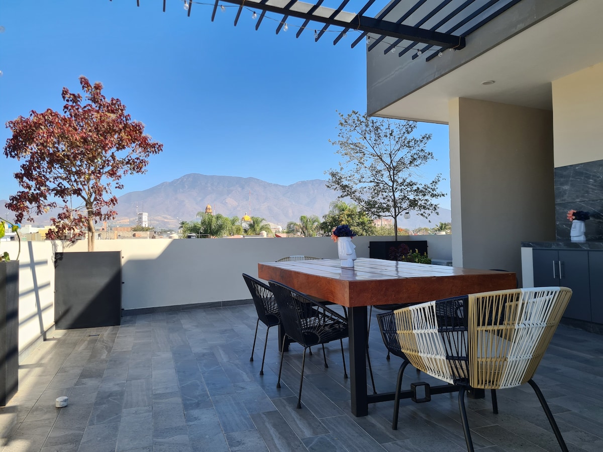 A rooftop terrace is presented, featuring a large wooden table surrounded by four black and gold woven chairs. Lush green trees and distant mountains create a scenic backdrop under a clear blue sky.