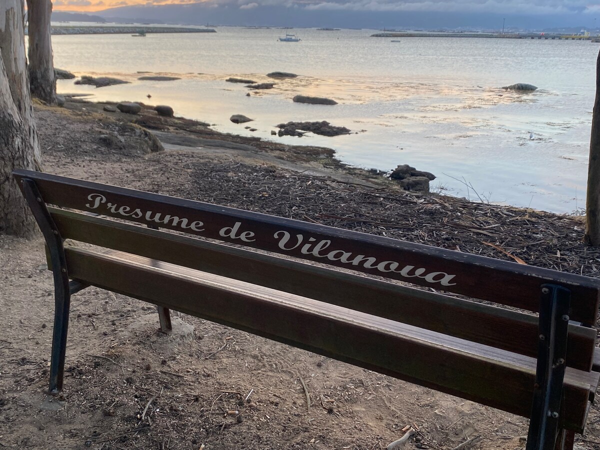 A wooden bench is positioned along the shoreline, featuring an inscription that reads 'Presume de Vilanova.' The tranquil water reflects the soft light of the setting sun, while rocky formations are visible in the foreground.