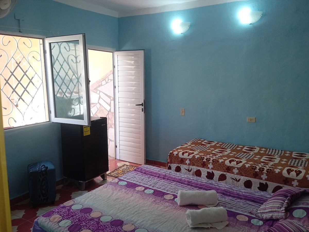 A light blue room features two beds with patterned bed linens, one with a vibrant design and the other in soft tones. A window offers natural light and ventilation, while a small refrigerator is visible against the wall. Fresh towels are neatly arranged on the beds.