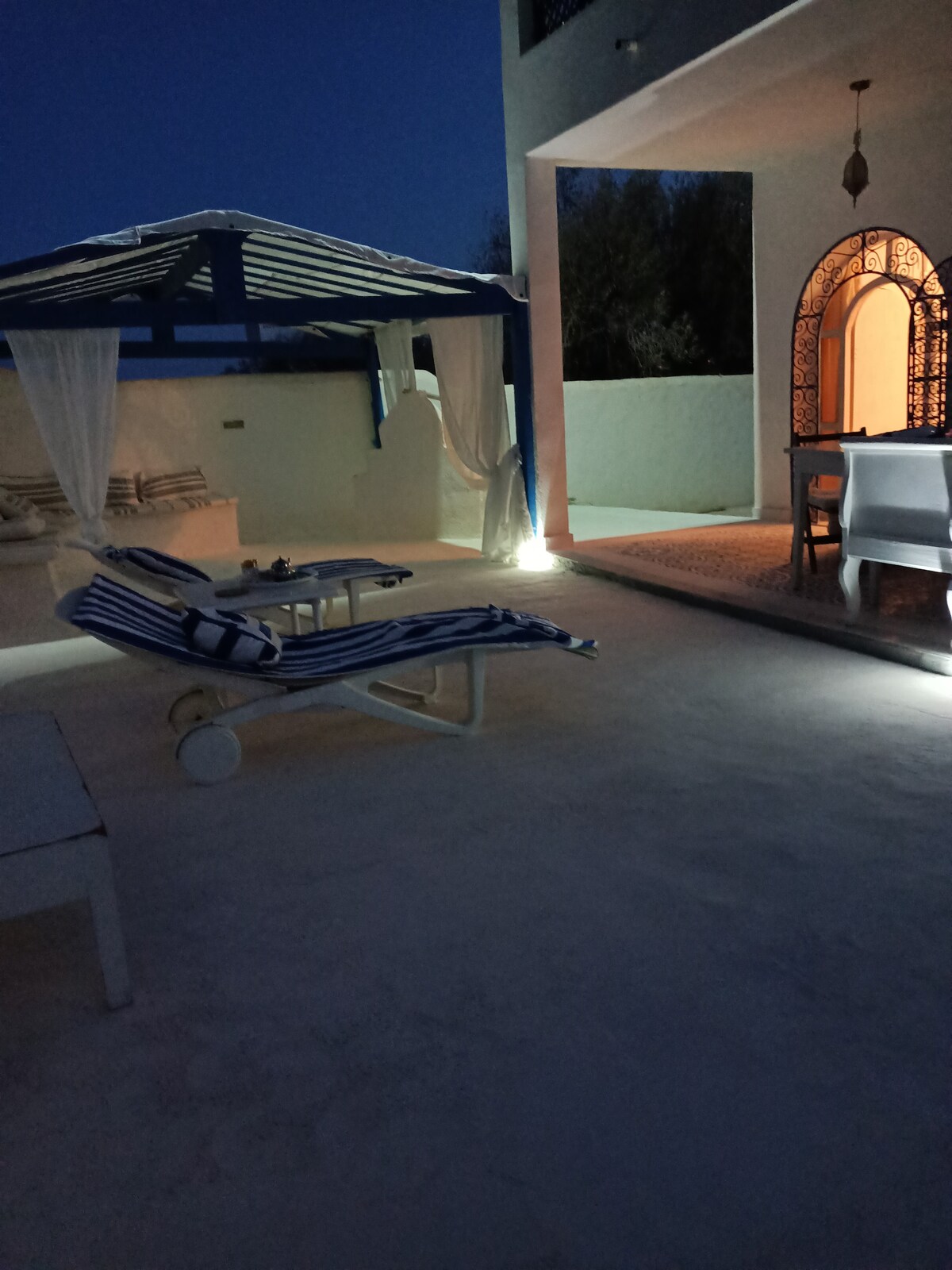 A tranquil outdoor area is illuminated softly at dusk, featuring a lounger with striped fabric. Nearby, a covered seating area provides shade, while a stylish dining set is visible through an arched doorway framed by intricate design elements.