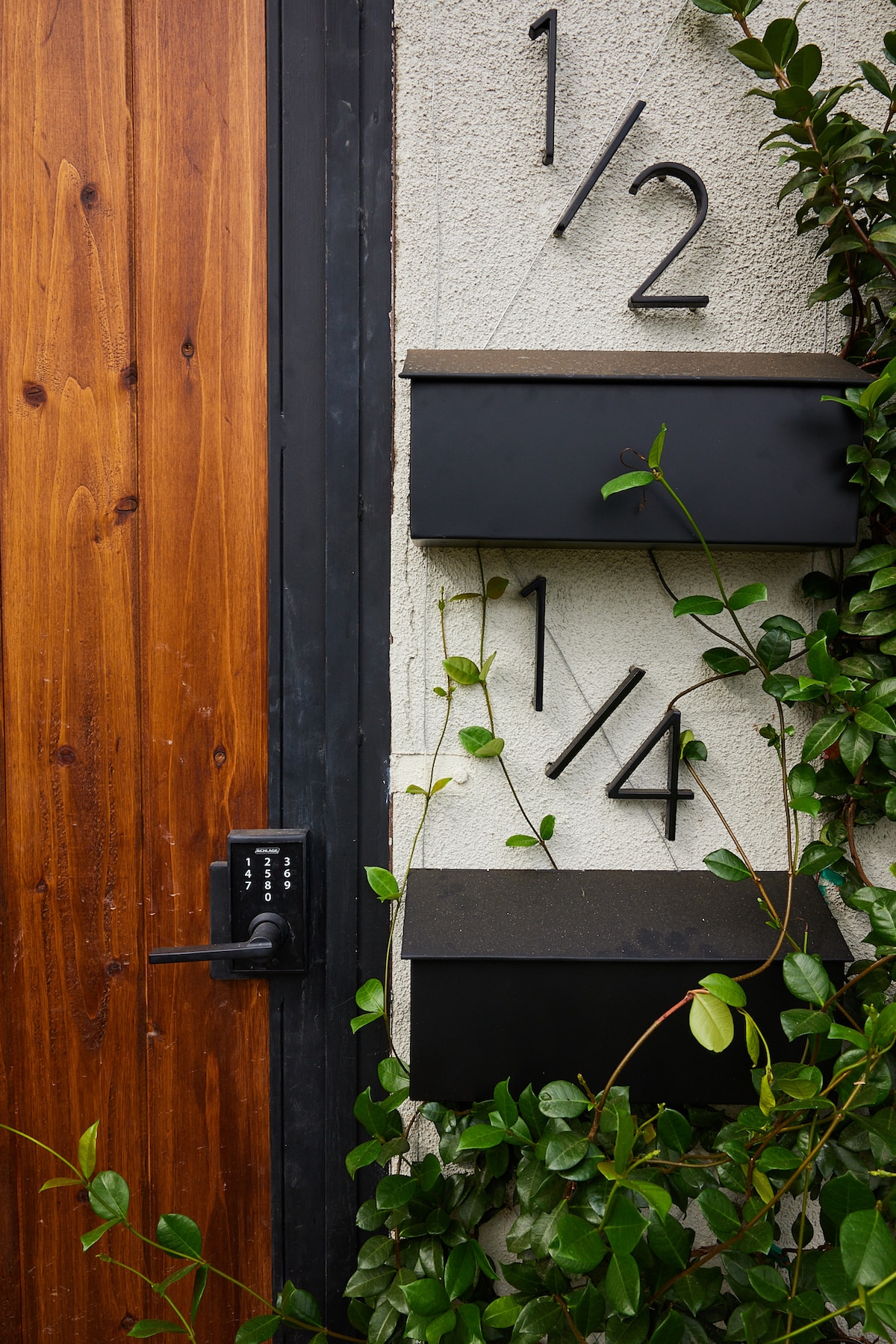 An entrance features a sturdy wooden door alongside a keypad lock, offering convenient self-check-in. Light green vines climb the exterior beside modern house numbers, indicating the unit's designation. The neutral walls provide a simple yet inviting appearance.