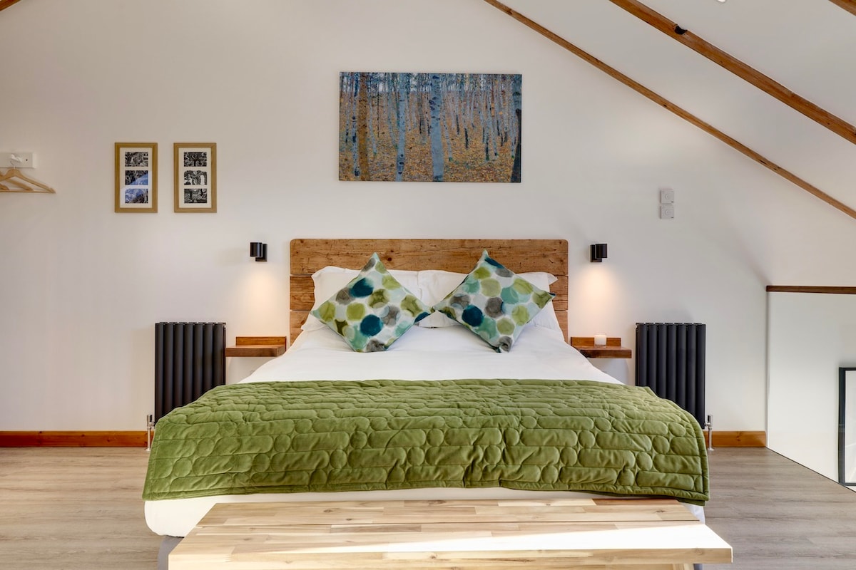 A well-appointed bedroom features a spacious bed topped with a textured green throw and decorative cushions. Artwork depicting trees adorns the wall above the bed. Two bedside lamps provide soft lighting, and a wooden bench sits at the foot of the bed, enhancing the room's design.
