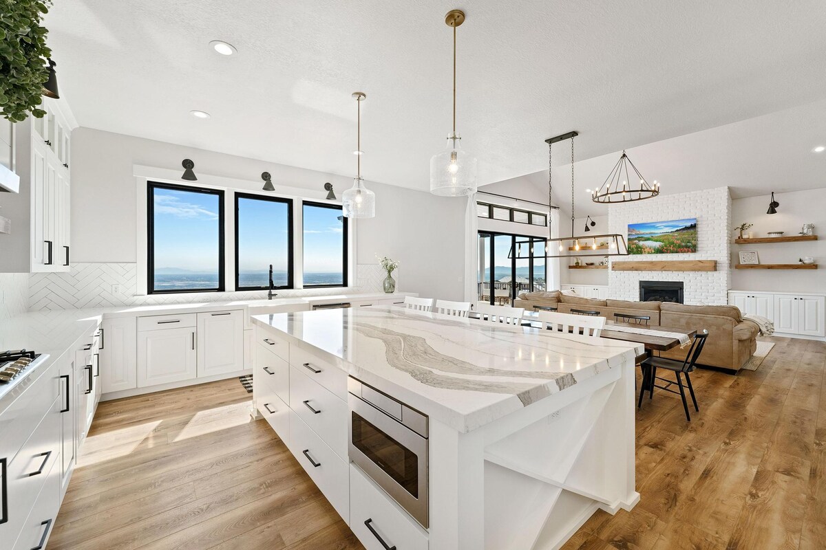 A modern kitchen features a large marbled island with bar seating, complemented by contemporary cabinetry. Natural light floods the space through wide windows that offer stunning views. Nearby, a cozy living area is furnished with a comfortable sectional and shelves displaying decorative items.
