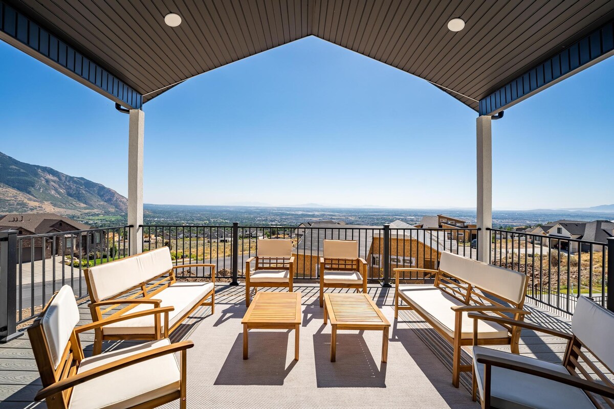 An outdoor seating area is showcased under a spacious roof, featuring several wooden chairs and tables arranged for relaxation. Panoramic views of the mountains and valley are visible through the open railing, providing a peaceful and inviting space for gatherings.