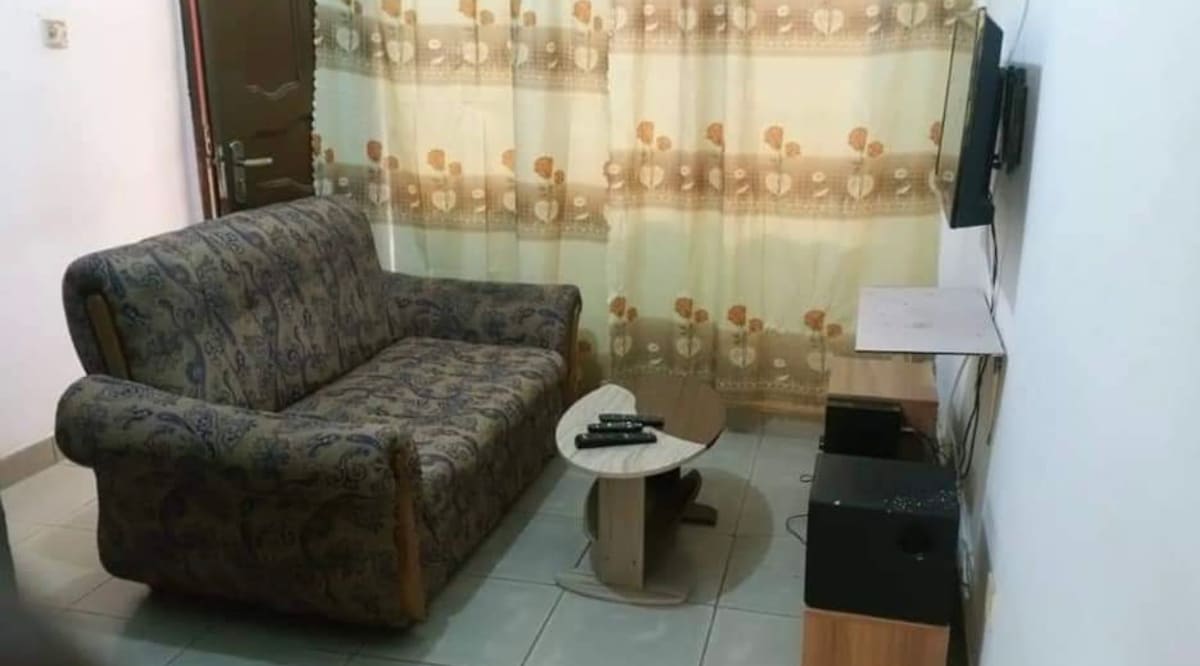 A living area features a patterned sofa paired with a small wooden coffee table. A media console is situated underneath a wall-mounted television, and sheer curtains adorn the window, allowing soft light to fill the room.