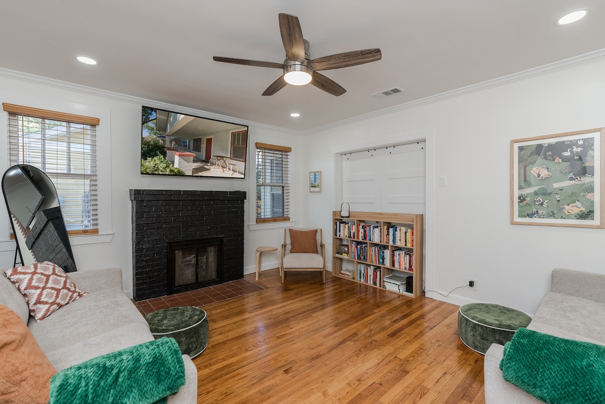 A spacious living area features two light-colored sofas and a wooden bookshelf filled with books. A ceiling fan is suspended above, and natural light filters in through the windows. A decorative green pouf and a neutral armchair add comfort to the space.