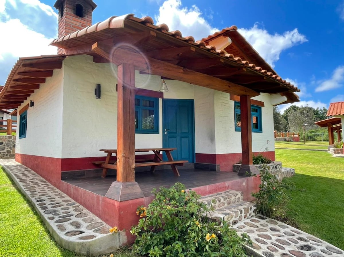 A charming cottage with a vibrant blue door is nestled under a tiled roof. The porch features a wooden picnic table and a stone pathway leads to the well-maintained lawn, surrounded by greenery and decorative flowers.