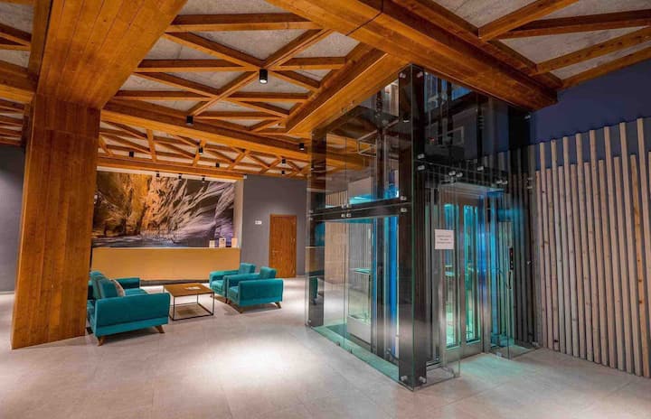 A modern lobby showcases a sleek glass elevator surrounded by comfortable teal seating. The ceiling features an intricate wooden structure, adding warmth. Natural stone artwork decorates the wall behind, enhancing the inviting ambiance of the space.