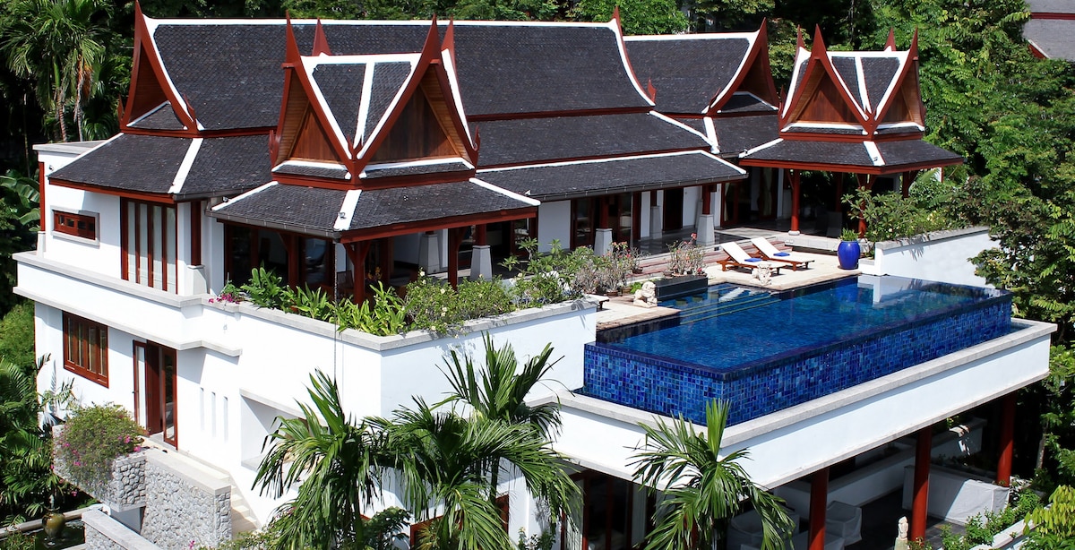 The villa's exterior showcases distinct Thai architectural elements, including intricate rooflines and wooden accents. A stunning infinity pool extends from the terrace, surrounded by lush greenery and modern outdoor furniture, creating an inviting space for relaxation.