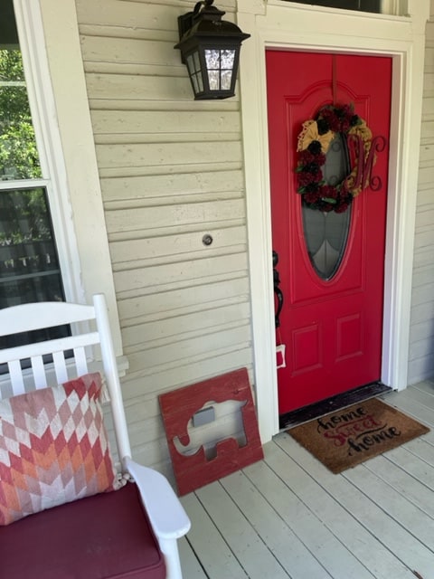 A welcoming front porch features a bright red door adorned with a decorative wreath. A vintage-style lantern casts a warm glow. A rocking chair is positioned beside a small elephant-shaped decoration, accompanied by a doormat that reads 'Home Sweet Home'.