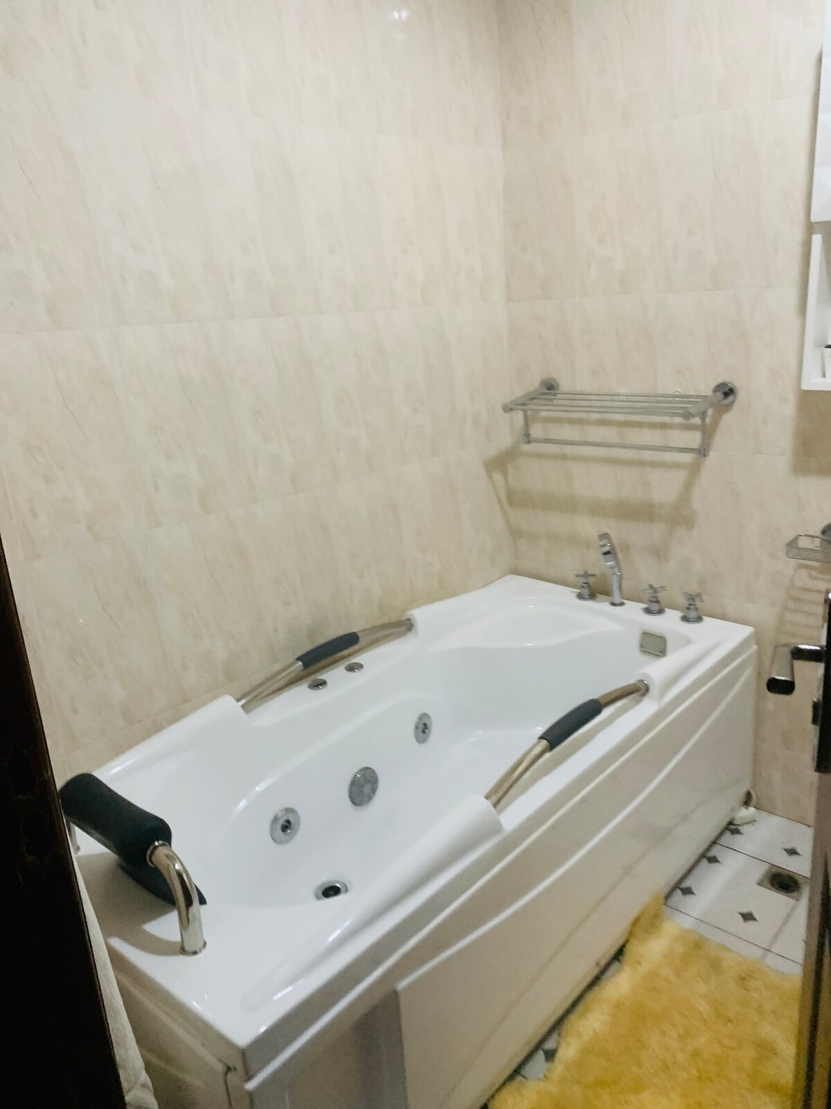A modern bathroom features a spacious whirlpool tub with adjustable jets. Light-colored tile walls and a textured floor mat create a clean and soothing atmosphere. A stainless steel towel rack is mounted above the tub, providing convenient storage.