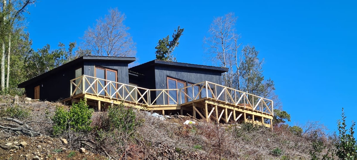 A modern house is positioned on a hillside, featuring a wooden deck that surrounds the structure. Large windows provide ample natural light, and the building contrasts with the surrounding trees. A clear blue sky fills the backdrop, enhancing the sense of openness.