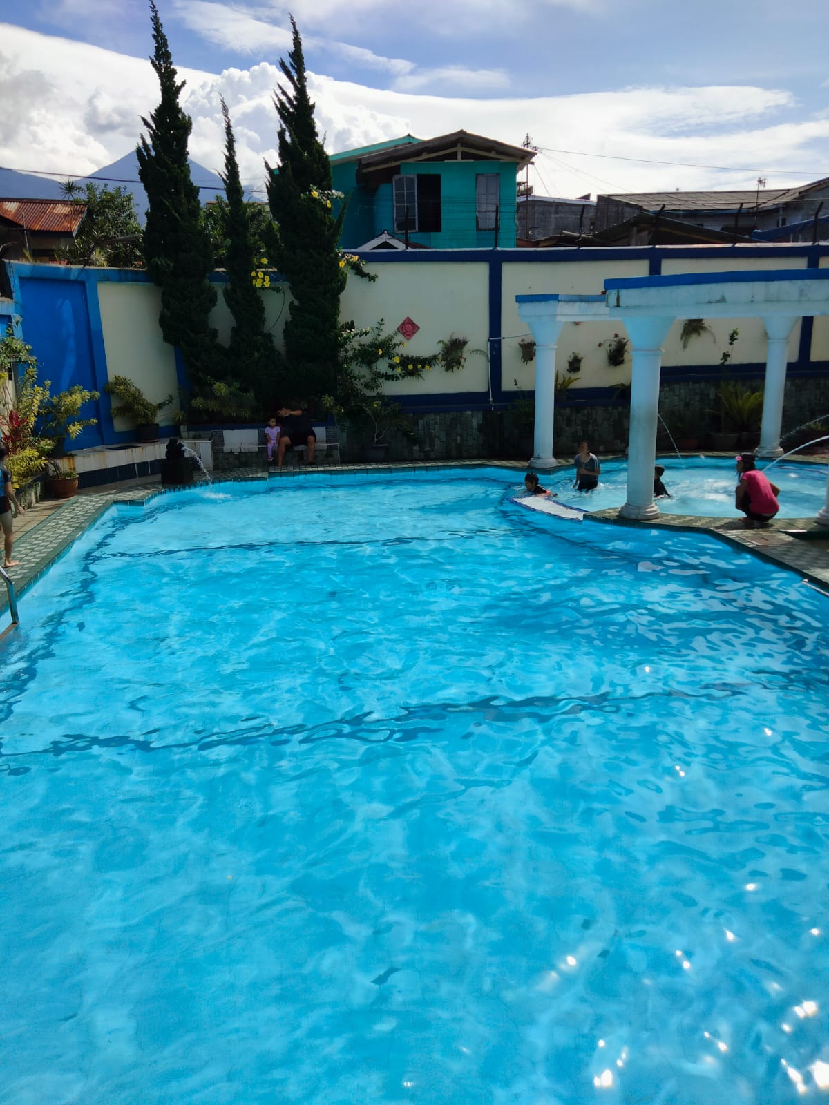 A serene swimming pool is featured, surrounded by lush greenery and tall, narrow trees. The clear blue water reflects sunlight, creating a shimmering effect. Steps lead into the pool, while a few individuals are seen enjoying the water.