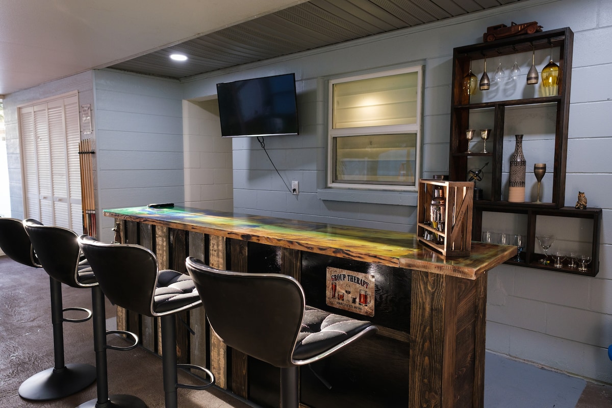 A sleek bar area is featured, with a wooden countertop and several stylish bar stools. A flat-screen television is mounted on the wall, and a display shelf showcases decorative glassware and bottles. The space is well-lit, enhancing its inviting atmosphere.