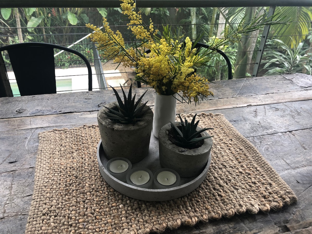 A wooden dining table is set with a textured woven placemat. Two concrete planters with small plants are paired with a slender white vase holding bright yellow flowers, while a circular tray with three tea light candles completes the arrangement, surrounded by lush greenery in the background.