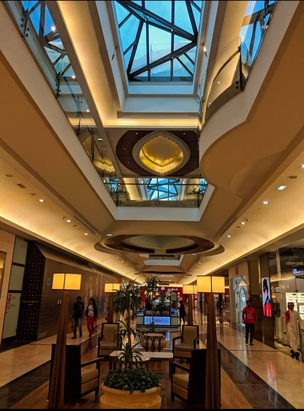 An elegantly designed corridor is illuminated by natural light streaming through a glass skylight. Modern lounge seating is arranged in a central area, flanked by two decorative lamps. Storefronts are visible in the background, providing a vibrant shopping atmosphere.