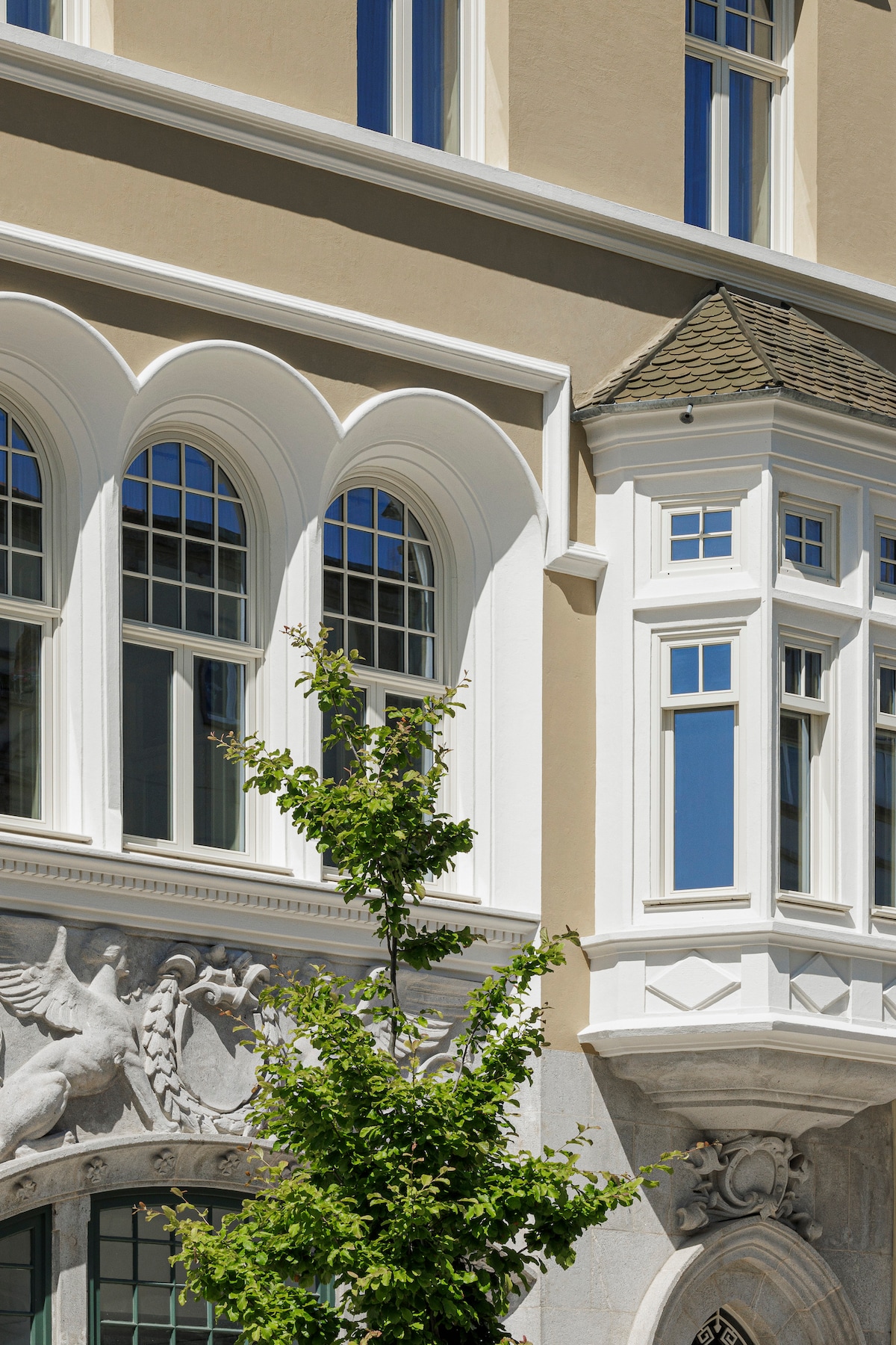 The exterior of a historic building is presented, showcasing architectural details such as arched windows and an ornate stone facade. A bay window is visible, framed by clean lines and elegant features, while a tree adds a touch of greenery to the scene.