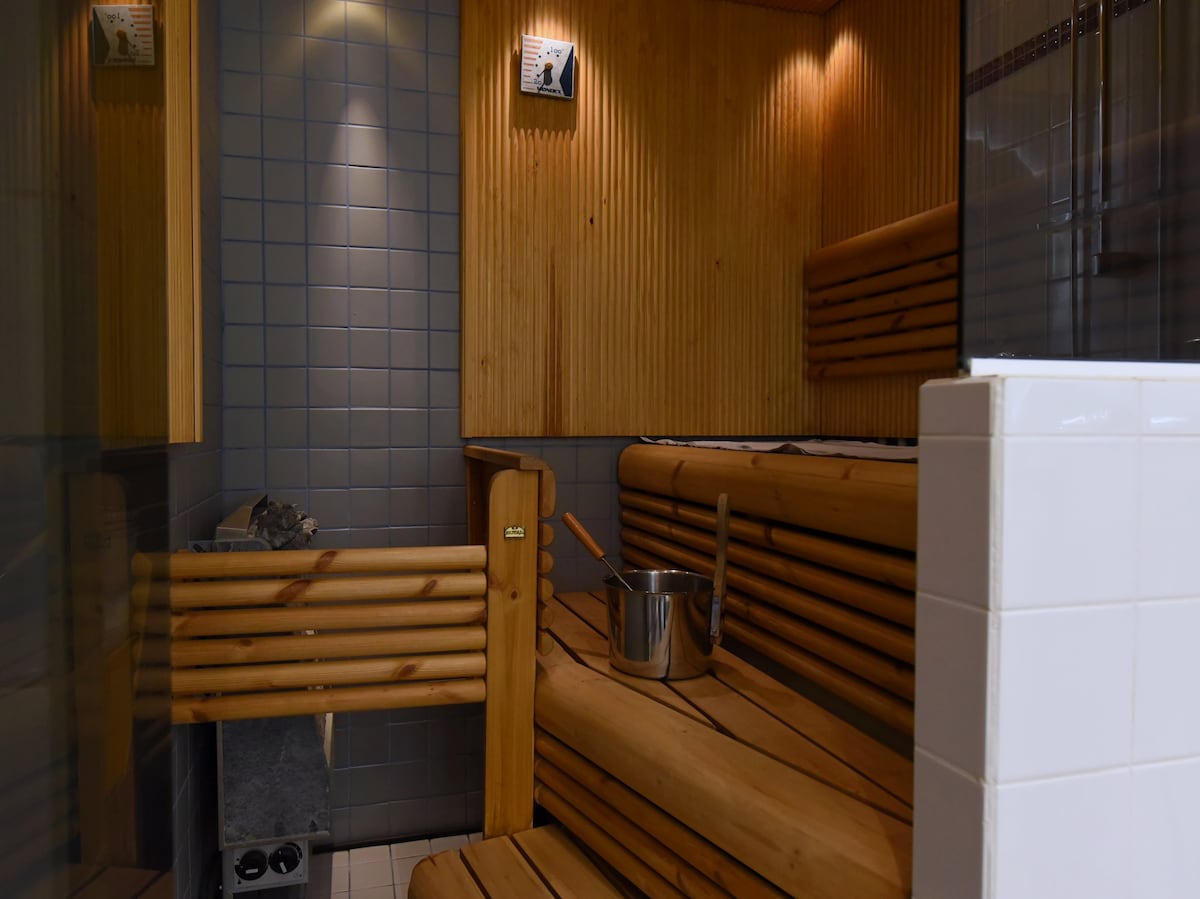 A sauna area features warm wooden benches and a stone wall, creating a calming environment. A bucket and ladle are positioned on the bench. Soft lighting highlights the natural wood finish, contributing to a serene atmosphere.