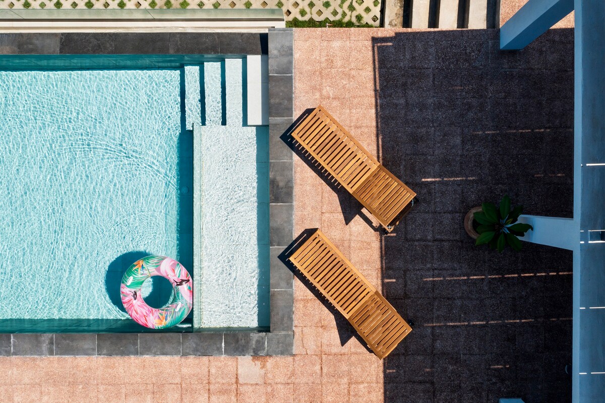 An aerial view of a modern swimming pool features a light blue surface and a colorful inflatable ring nearby. Two wooden sun loungers are positioned beside the pool area on a textured patio, offering a serene atmosphere for relaxation.