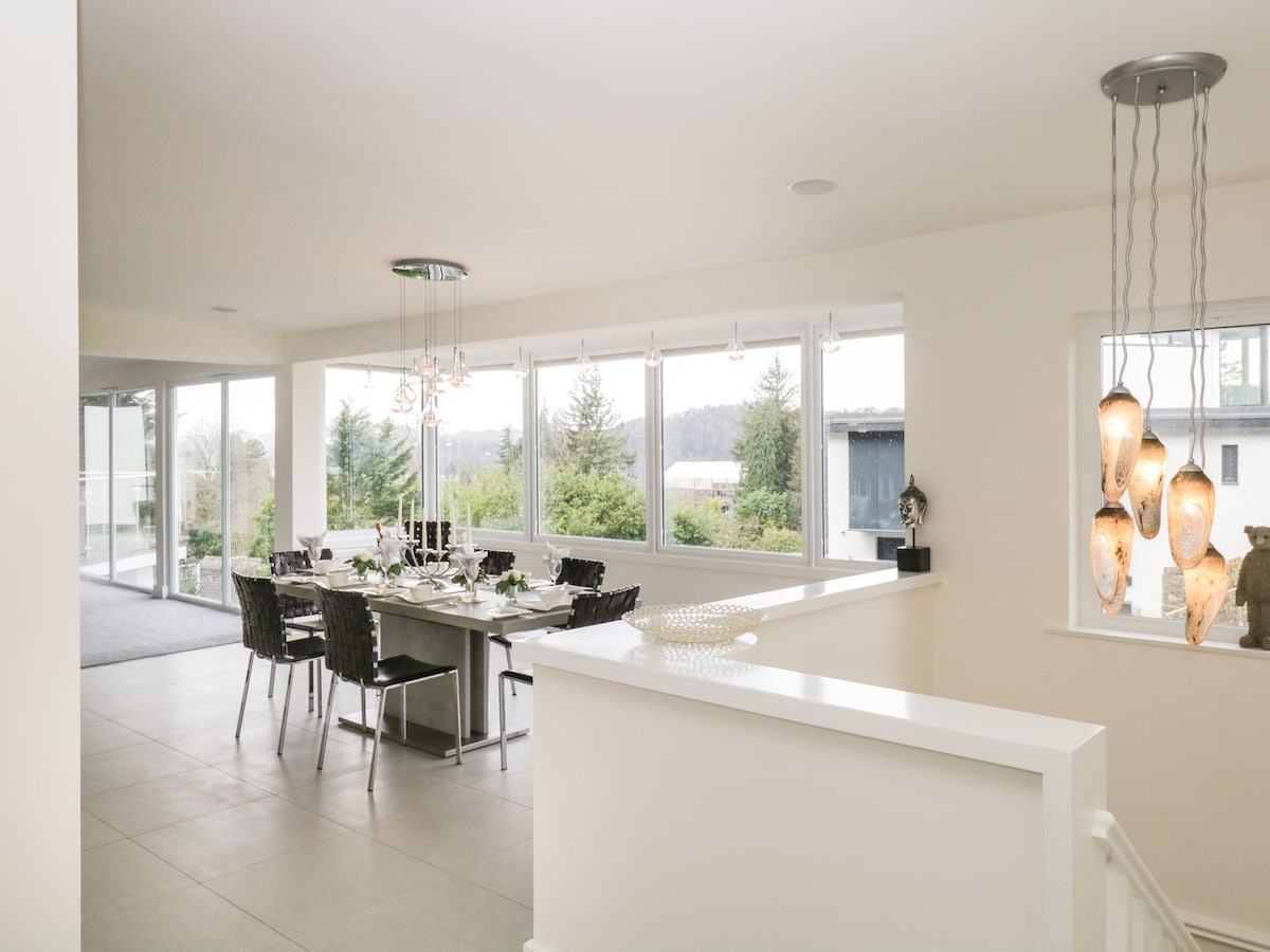 An airy dining area is highlighted by large windows letting in natural light, showcasing views of lush greenery. A modern dining table is set for meals, surrounded by contemporary dark chairs. Elegant pendant lighting features a mix of soft, warm tones, enhancing the inviting ambiance.