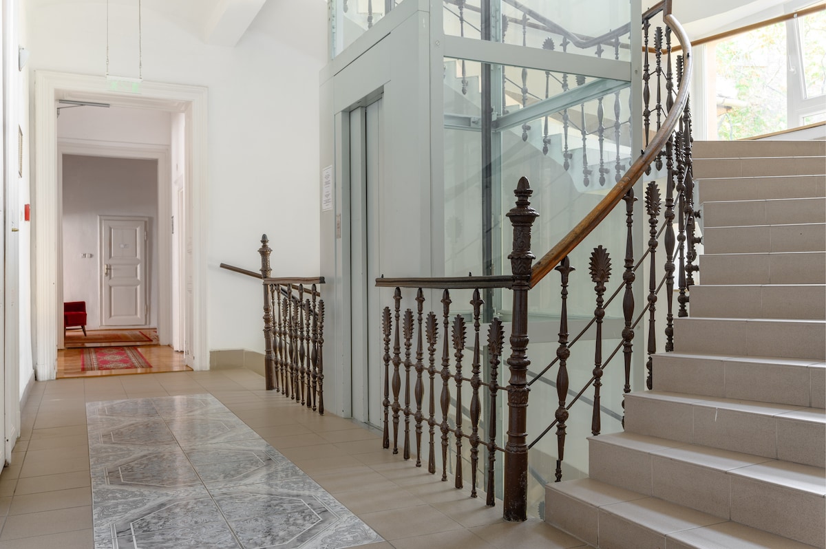 The hallway features an elegant staircase with a decorative wrought iron railing. An elevator is visible nearby, and the floor is covered with large tiles. Natural light streams through windows along the corridor, highlighting the minimalist design and soft color palette.