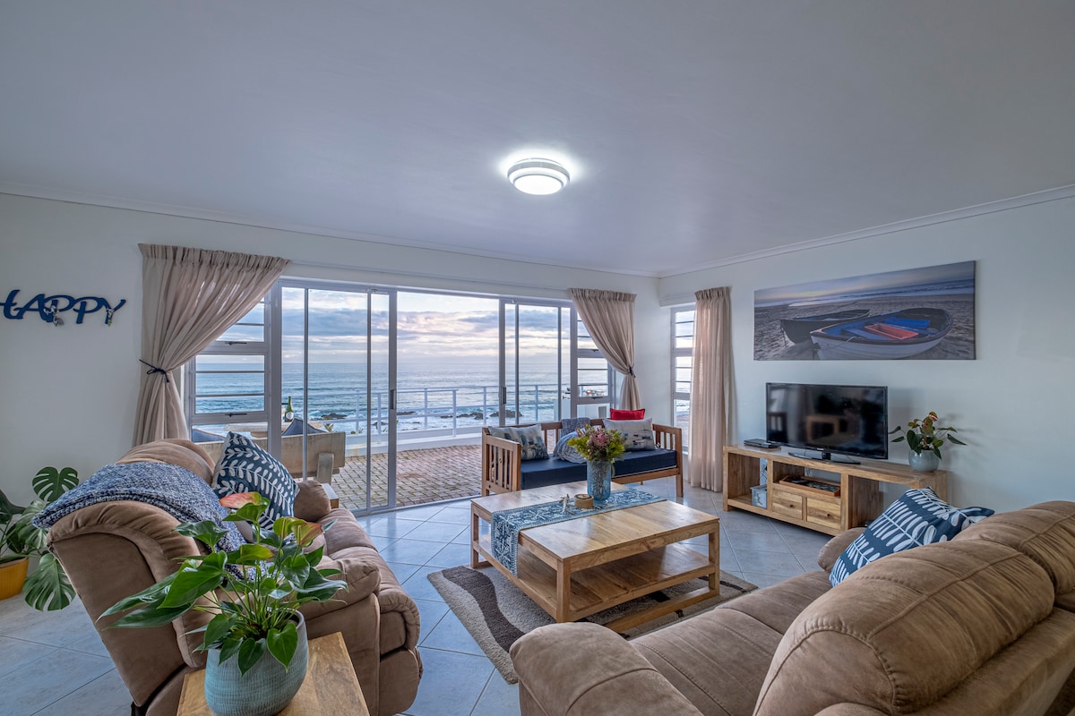 A spacious lounge area features large glass doors allowing natural light and showcasing views of the beach and ocean. Comfortable seating is provided by plush sofas, complemented by a wooden coffee table and side tables. A TV is positioned on a wooden unit, enhancing the inviting ambiance.