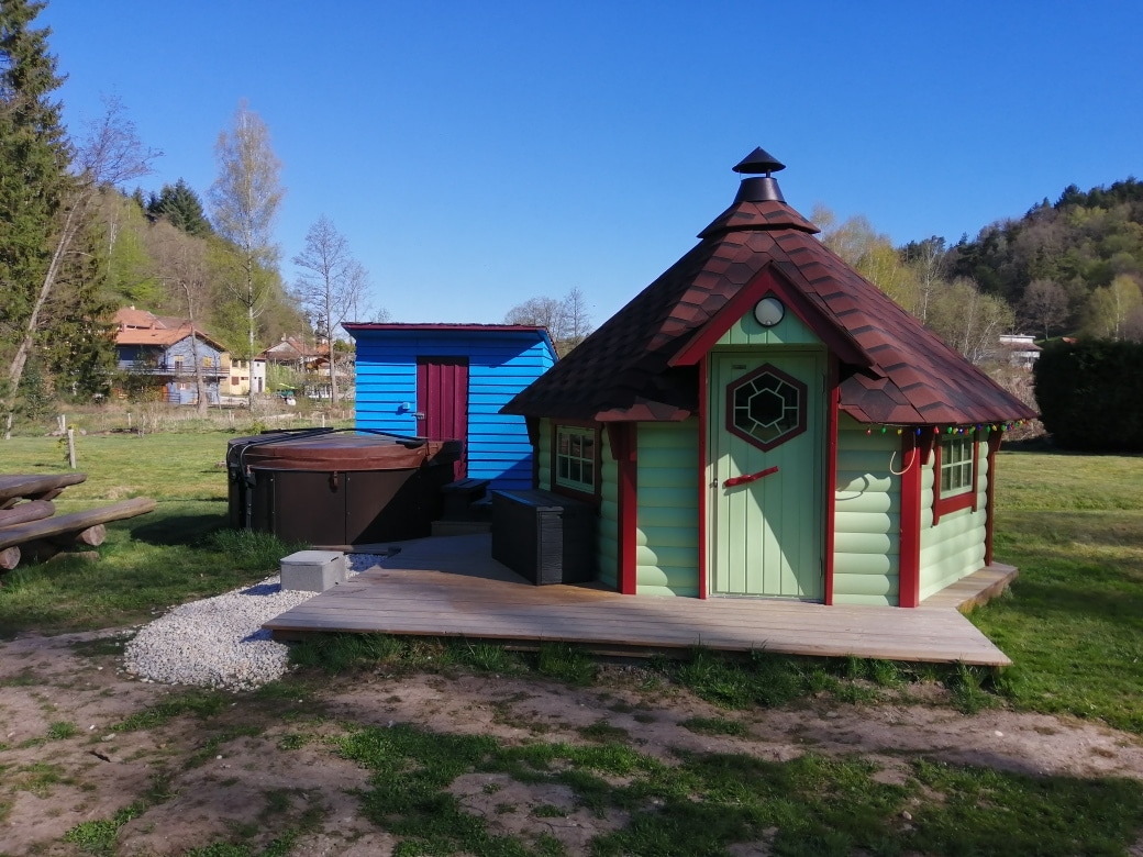 A quaint outdoor area features two colorful cabins, one in green with a unique roof design and detailed door, and another in blue. A wooden deck is showcased with a small hot tub nearby, surrounded by grass and gravel. Trees and distant buildings are visible in the background.