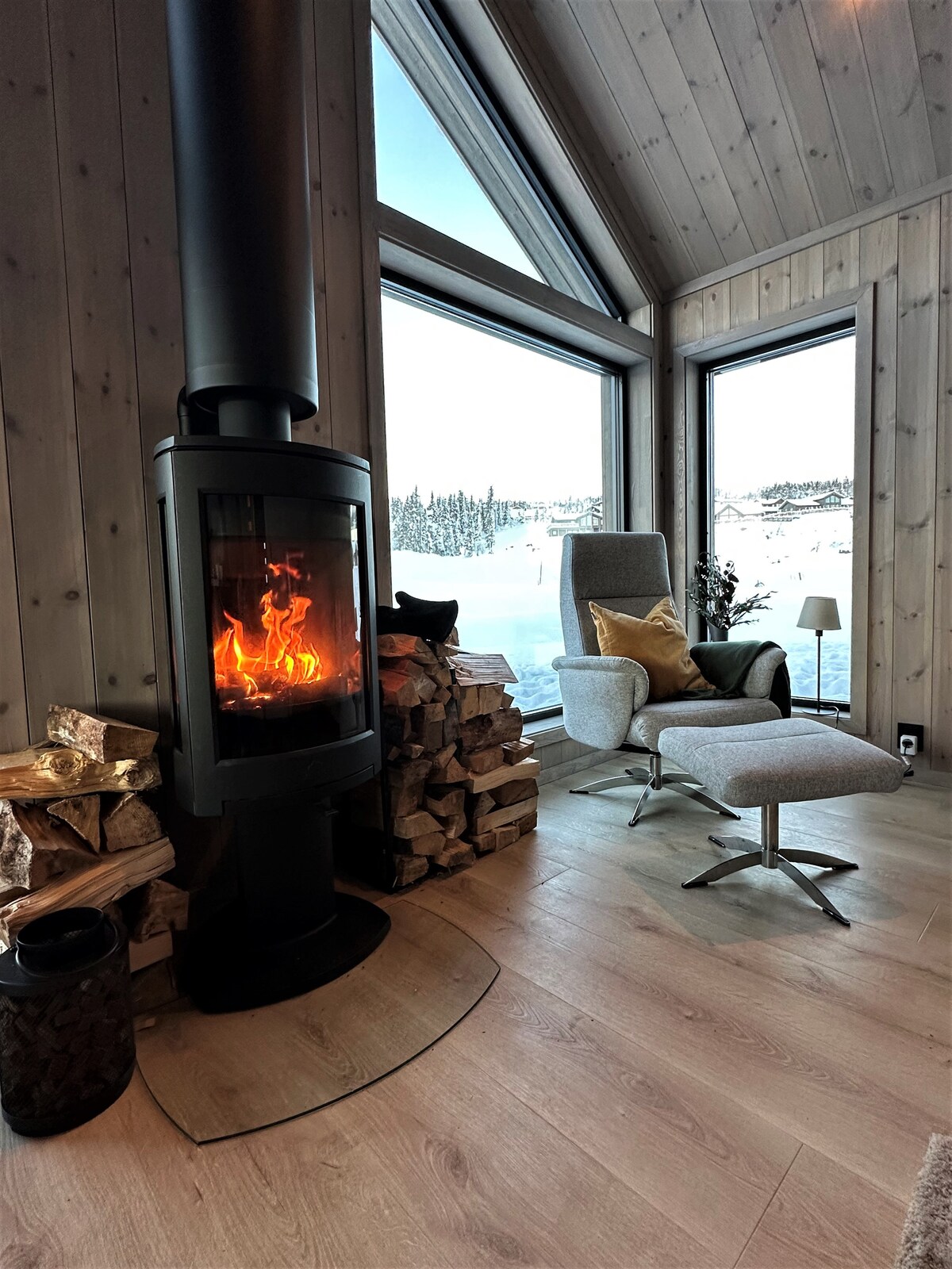 A modern seating area is complemented by a sleek armchair and ottoman, positioned beside a contemporary fireplace. The fireplace casts a warm glow, while large windows offer a view of the winter landscape outside, framed by wood-paneled walls.
