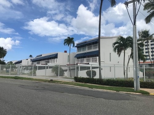 A row of white villas is seen behind a wrought iron fence, showcasing a combination of balconies and large windows. Lush greenery lines the property, with palm trees providing shade. The sky features scattered clouds, contributing to a bright atmosphere.