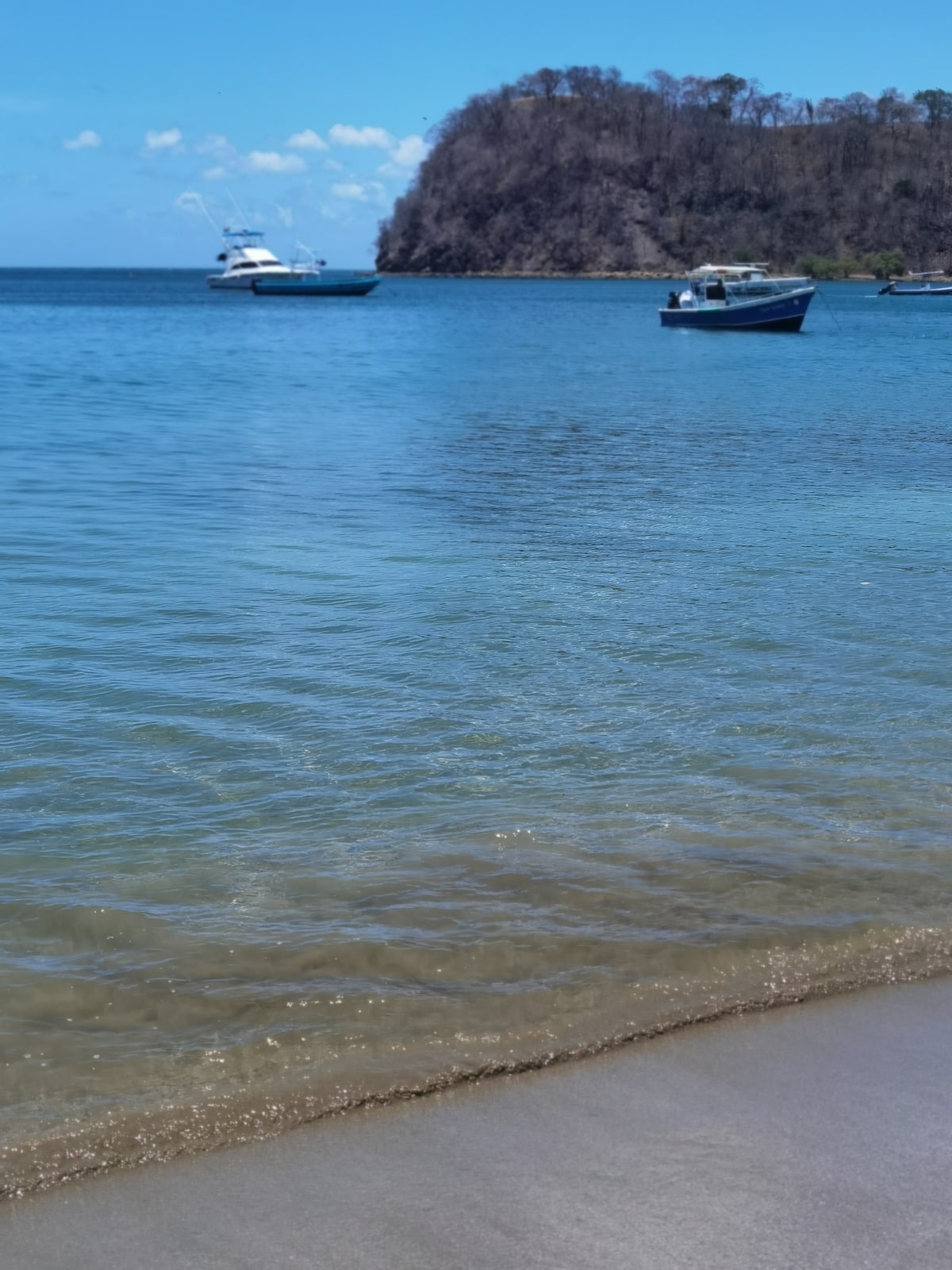 A calm beach scene is depicted, showcasing clear blue water that gently waves onto a sandy shore. Several boats are anchored in the distance, while rocky formations are visible along the coastline under a bright sky.