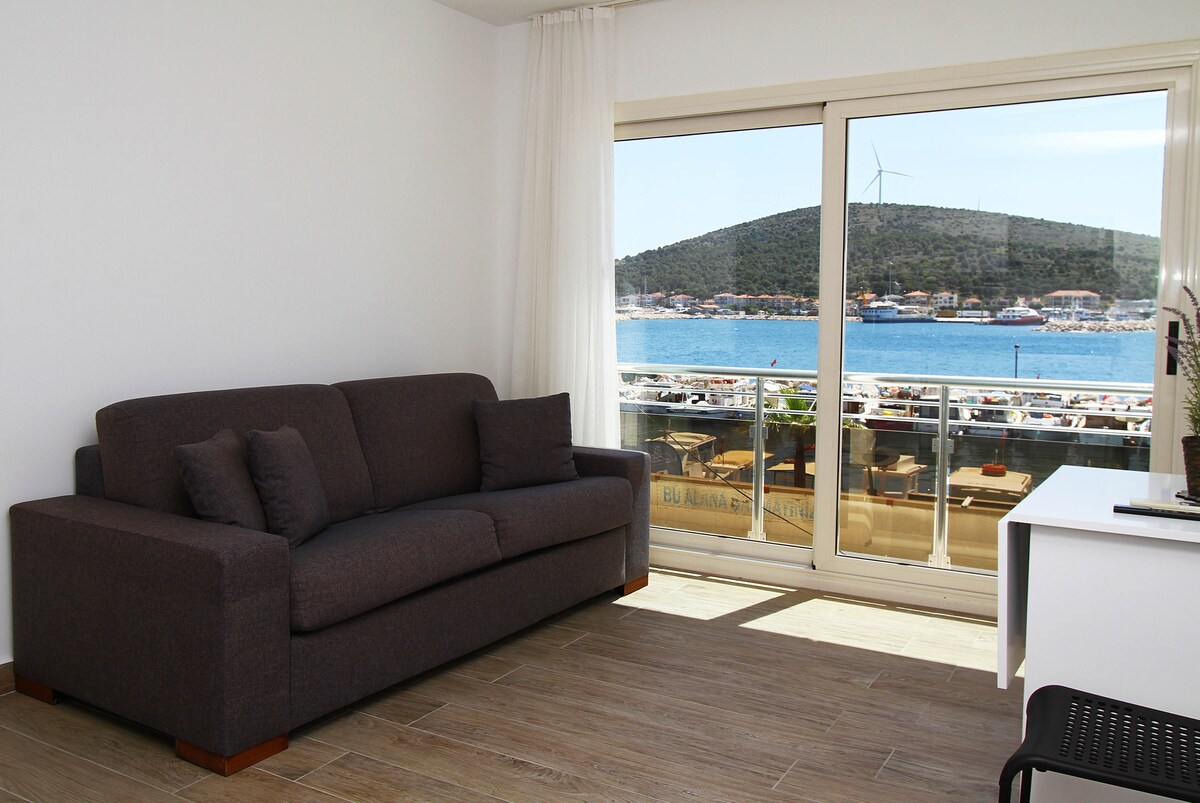 A comfortable dark sofa is positioned beside a large window, offering a view of the waterfront and nearby hills. Natural light fills the space, enhancing the neutral color palette of the room. A modern desk is visible, adding functionality to the area.