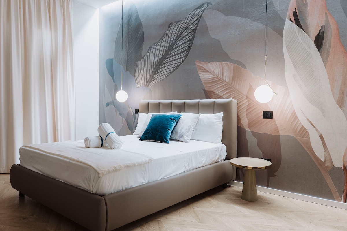 A spacious bedroom is depicted, featuring a large bed with neatly arranged white linens and a blue accent pillow. A subtle leaf-patterned mural adorns the wall, while two hanging lights cast a soft glow. A small, round side table complements the modern decor.