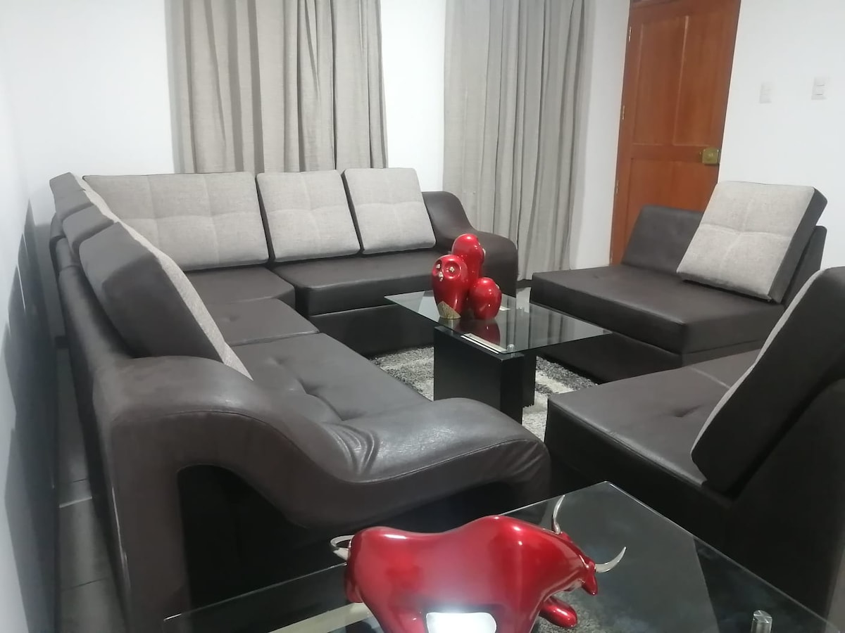 A spacious seating area is furnished with a large sectional sofa and a matching armchair, arranged around a glass coffee table. Soft grey curtains frame the windows, and decorative red figures are placed on the table, adding a touch of color to the neutral-toned furnishings.
