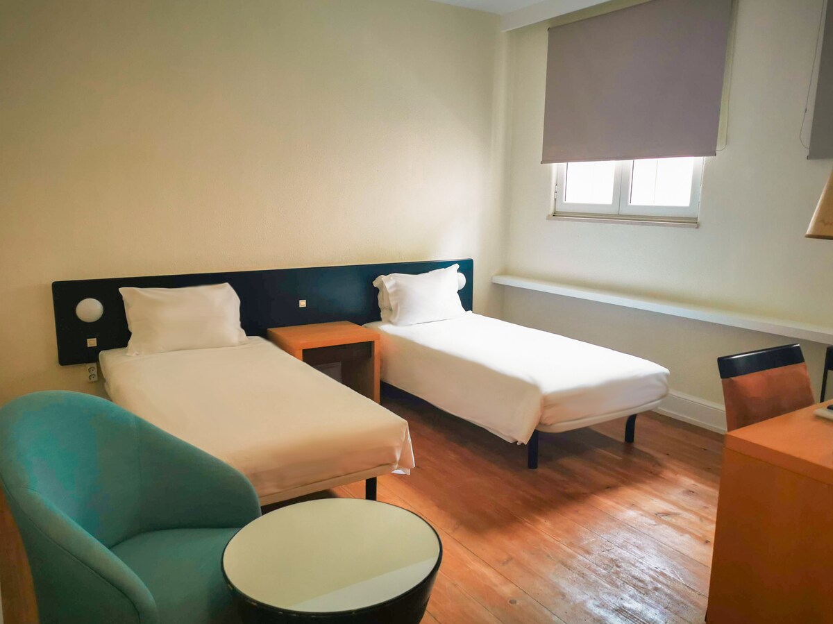 A twin room features two spacious beds with white linens, arranged next to a simple wooden nightstand. A small round table and a light blue armchair are positioned nearby, with a wooden desk visible in the room. Natural light enters through a window with blinds.