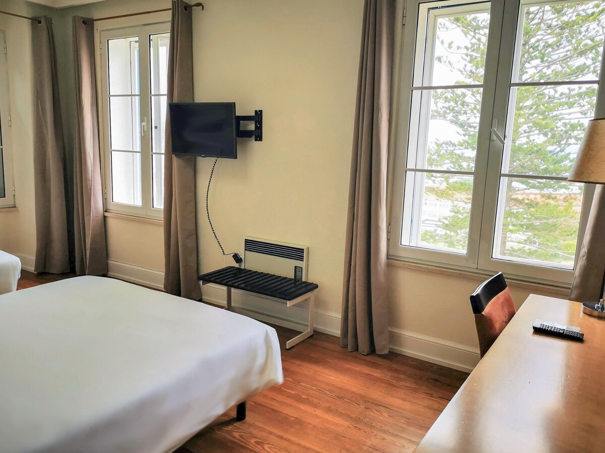 The private room features a spacious bed with crisp white linens, a wall-mounted TV, and a wooden desk. Natural light filters through two large windows adorned with light curtains, offering a view of greenery outside. A shelf is positioned near the bed for convenience.