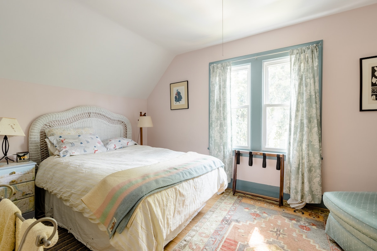 A serene bedroom features a large bed with a patterned blanket and decorative pillows. Natural light filters through two tall windows, adorned with patterned curtains. A small table sits beside the bed, while an area rug adds warmth to the hardwood floor.
