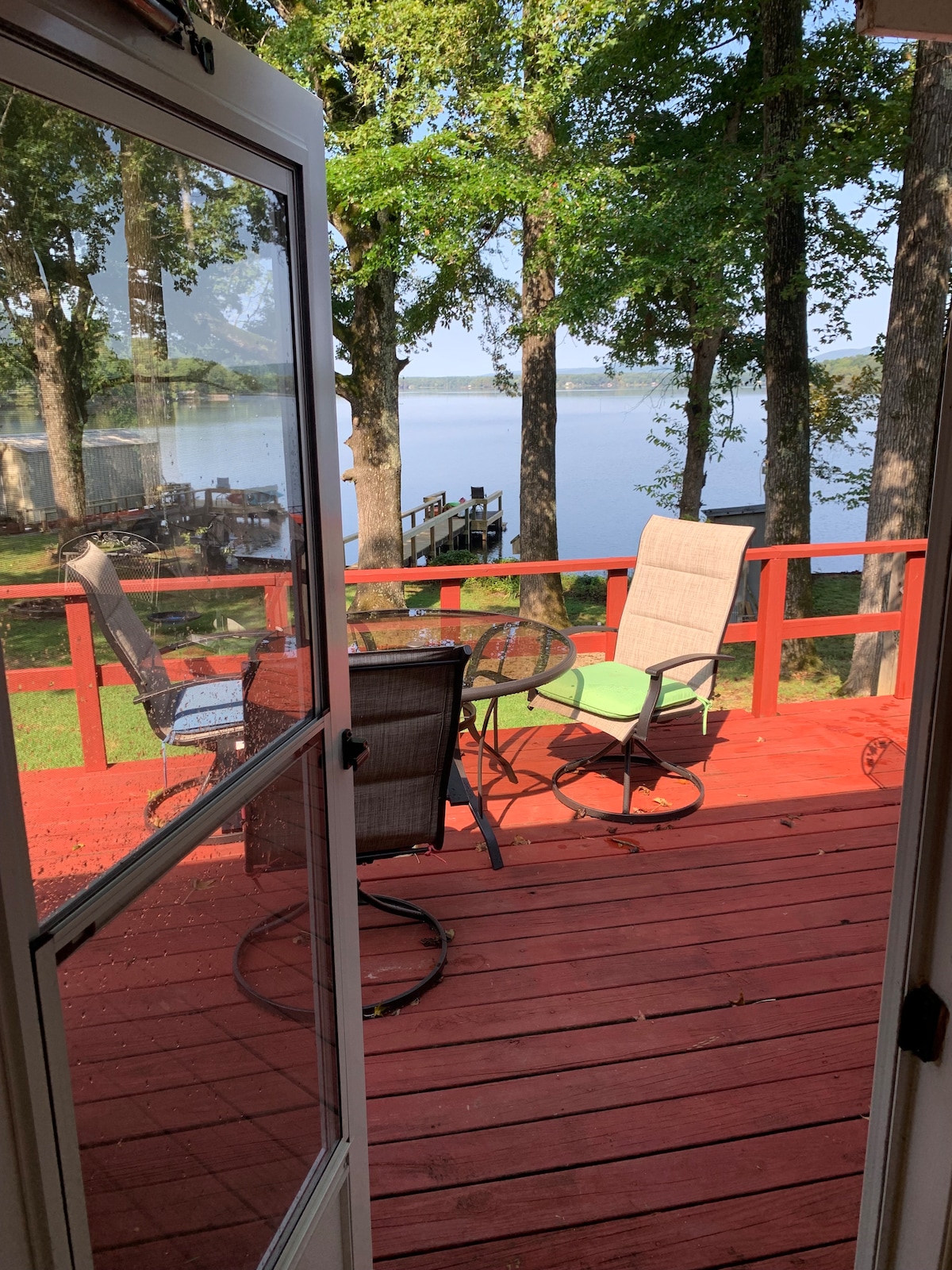 A view from the cabin door opens onto a red deck, furnished with a table and chairs. The serene lake is visible beyond the railing, surrounded by trees. A glimpse of the dock can be seen in the distance along with the calm water.