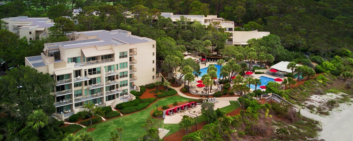 An aerial view showcases the resort property surrounded by lush greenery. The outdoor pool area is visible, along with lounge chairs and shaded seating. Paths weave through the landscaped grounds, leading towards the beach area that is partially visible in the foreground.