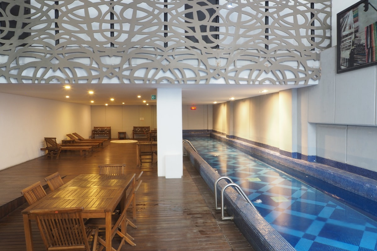 An indoor pool area features a long, tiled swimming pool with blue patterns. Wooden lounge chairs are arranged alongside the pool, and a dining set is positioned nearby, composed of a square table and matching chairs. Ambient lighting creates a relaxed atmosphere.