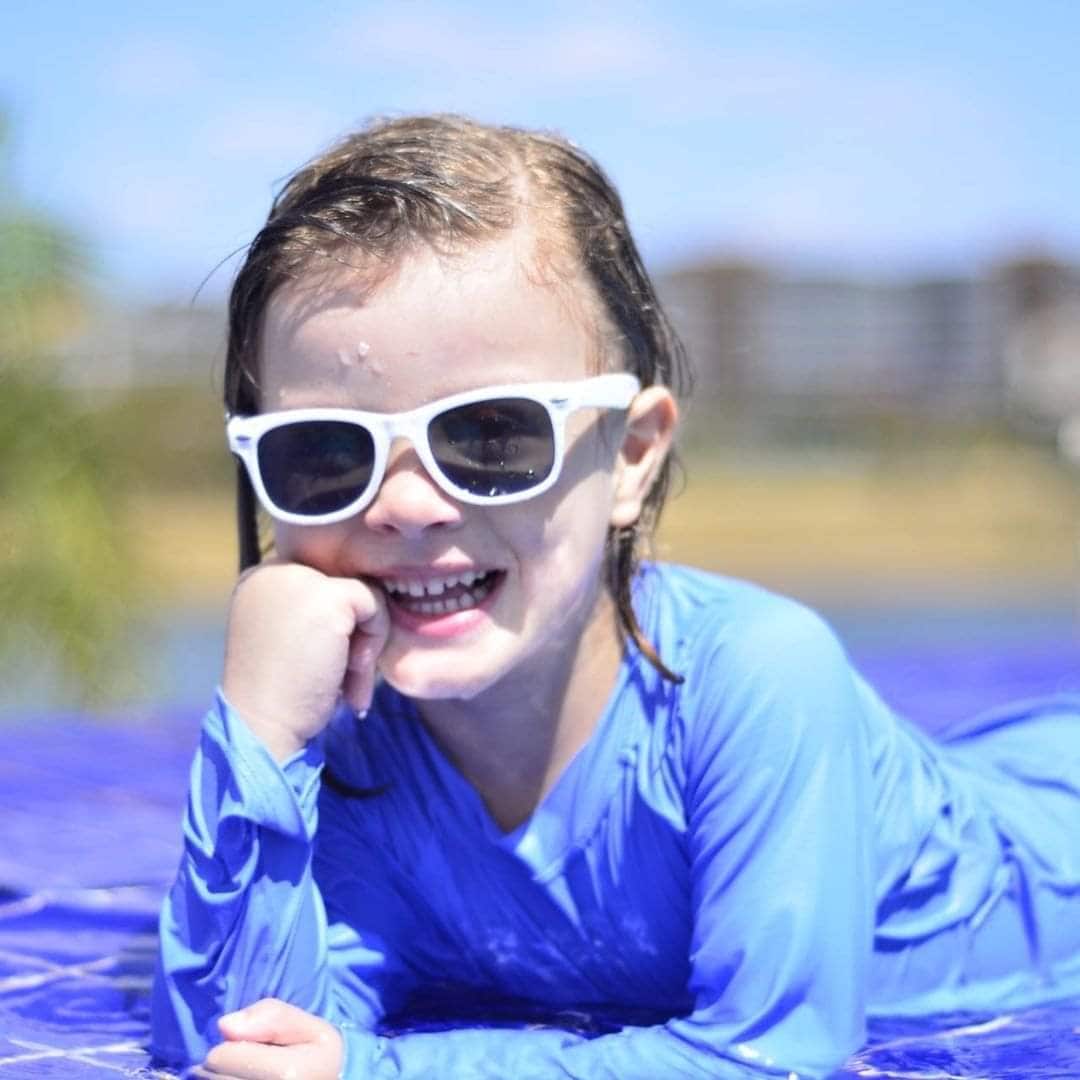 A child is seen lounging on a water mat, wearing a blue long-sleeve swim top and protective goggles. The calm blue water surrounds them, suggesting a warm and relaxing environment. In the background, buildings and vegetation blend into a serene lakeside scene.