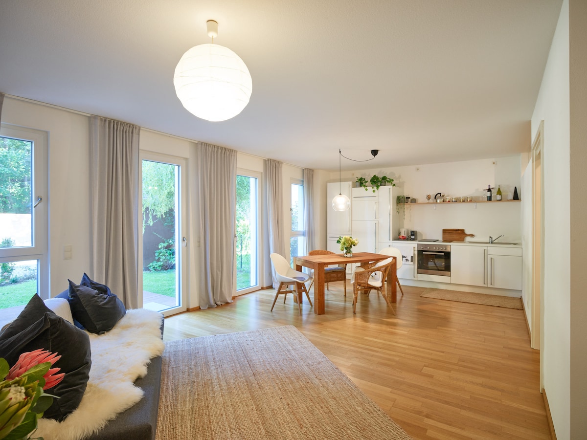 An open living area features a light wood floor and large windows allowing natural light. A round dining table is surrounded by white chairs and complemented by a small vase of flowers. A cozy sofa adorned with cushions is positioned nearby, and a kitchen area is visible in the background.