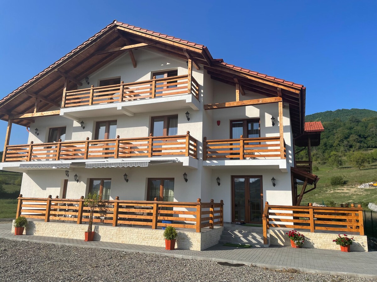 A charming three-story building with a rustic wooden facade is surrounded by landscaped grounds. Balconies with wooden railings provide views of the surrounding landscape. Bright flower pots line the entrance, creating an inviting approach. A clear blue sky tops the scene.