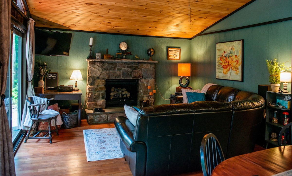 A cozy living area features a stone fireplace as a central focal point, surrounded by soft lighting from nearby lamps. Dark seating is complemented by warm wooden accents and a colorful piece of wall art. Large glass doors provide views of the outdoors.